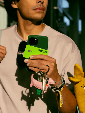 A man is walking down a shopping street. He holds his phone, keys, sunglasses and a Wise card in his hand.